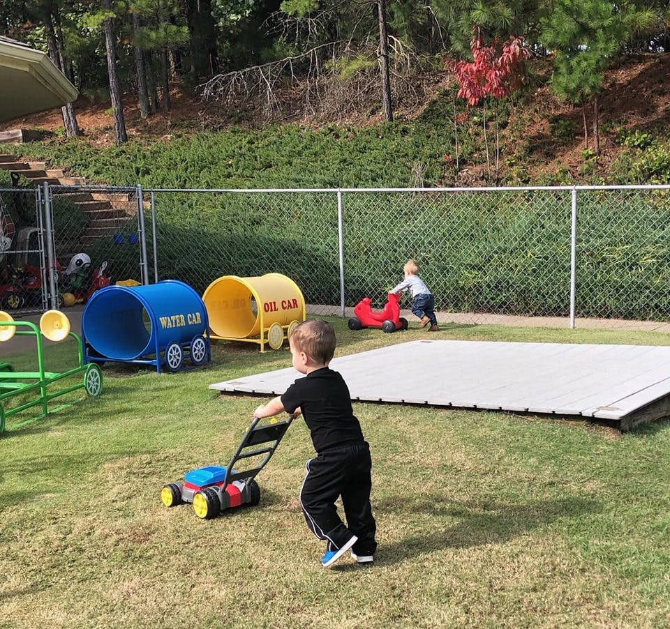 Child pushing a toy lawnmower on grass, another child on a ride-on toy