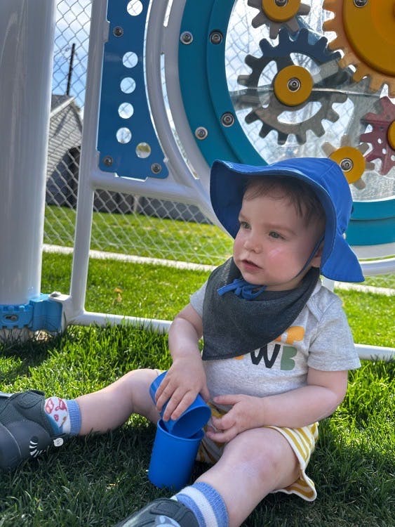 Child in a blue hat and bib playing with blue cups on grass