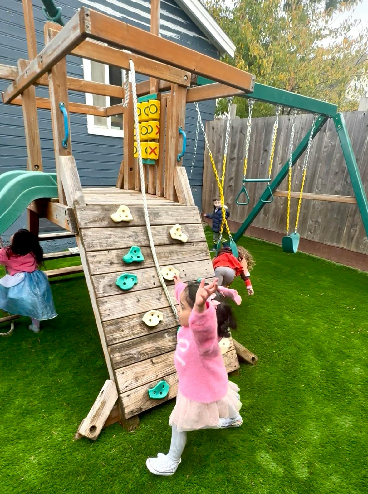 Children playing on a wooden playset with a slide, climbing wall, and swings