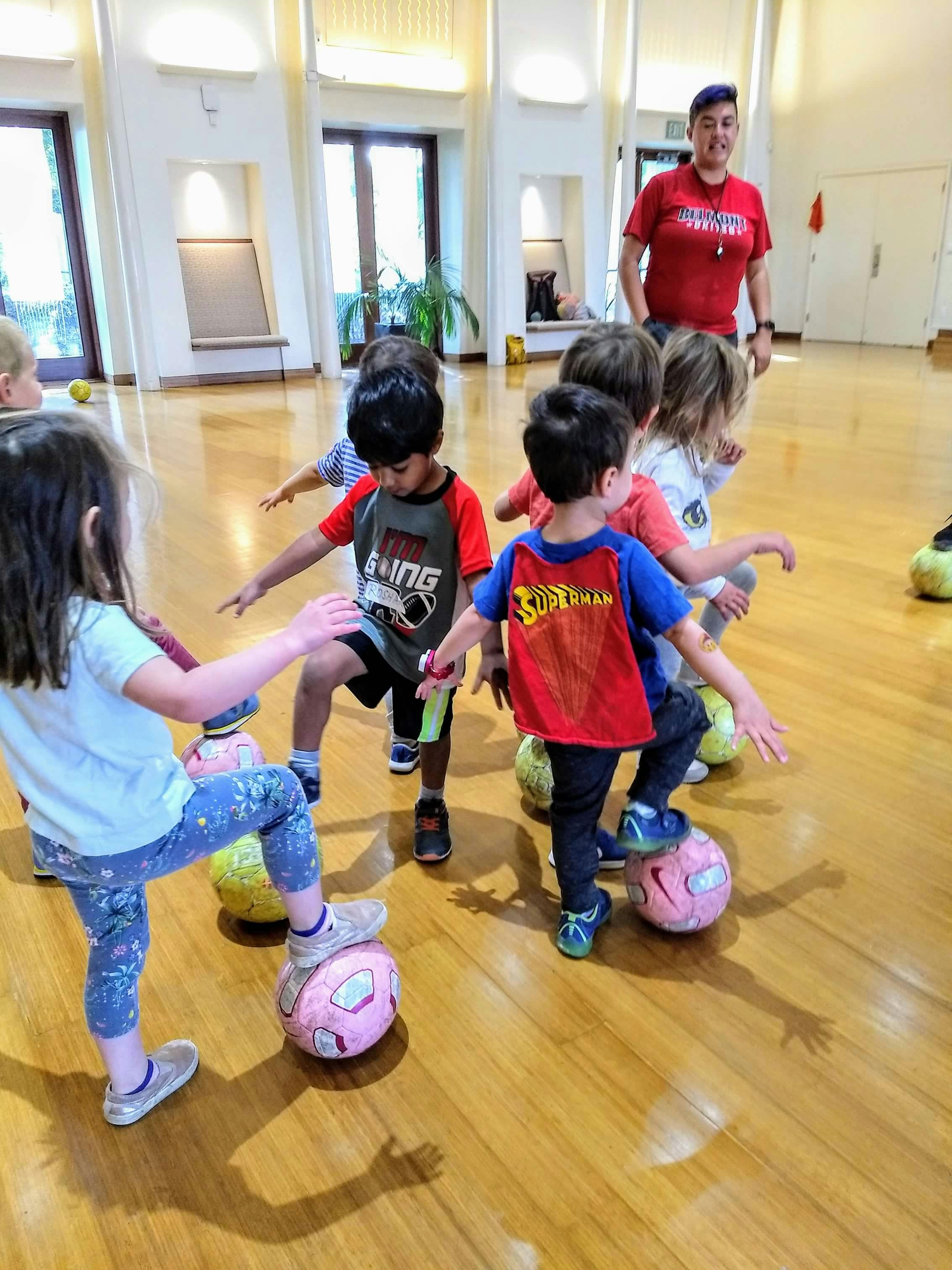 Children standing with feet on soccer balls in a room with an adult supervising