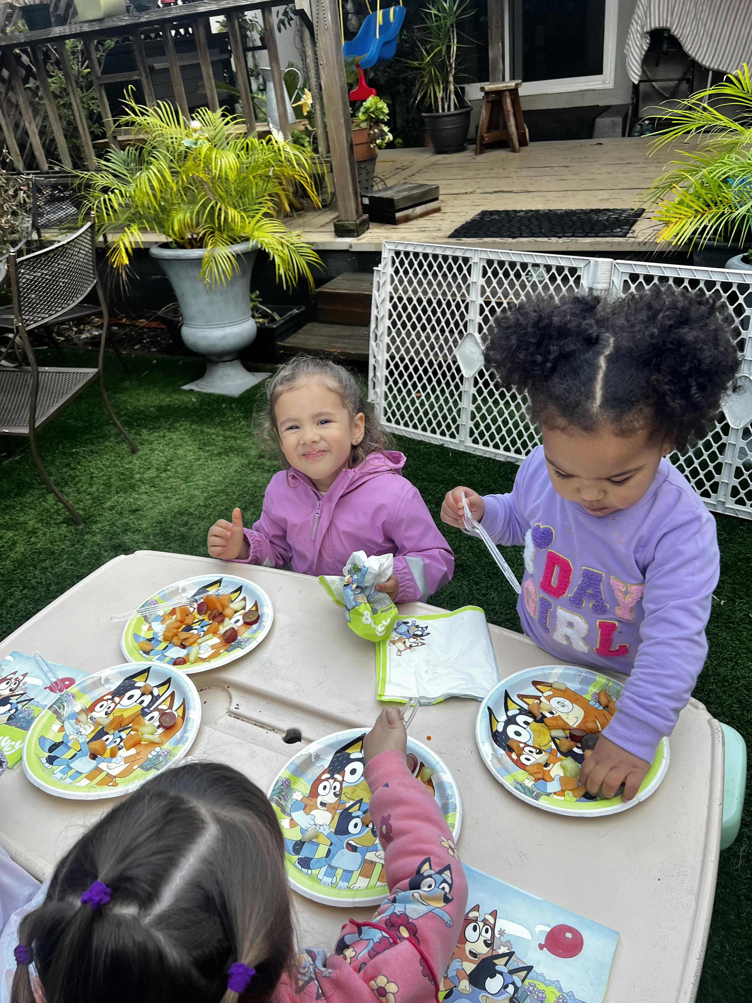 Three children seated at a table outdoors, eating from plates of fruit