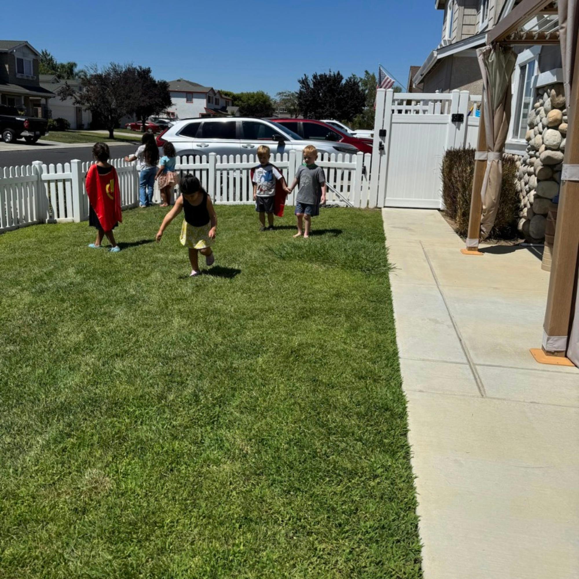 Children in capes playing on a grassy lawn next to a white fence