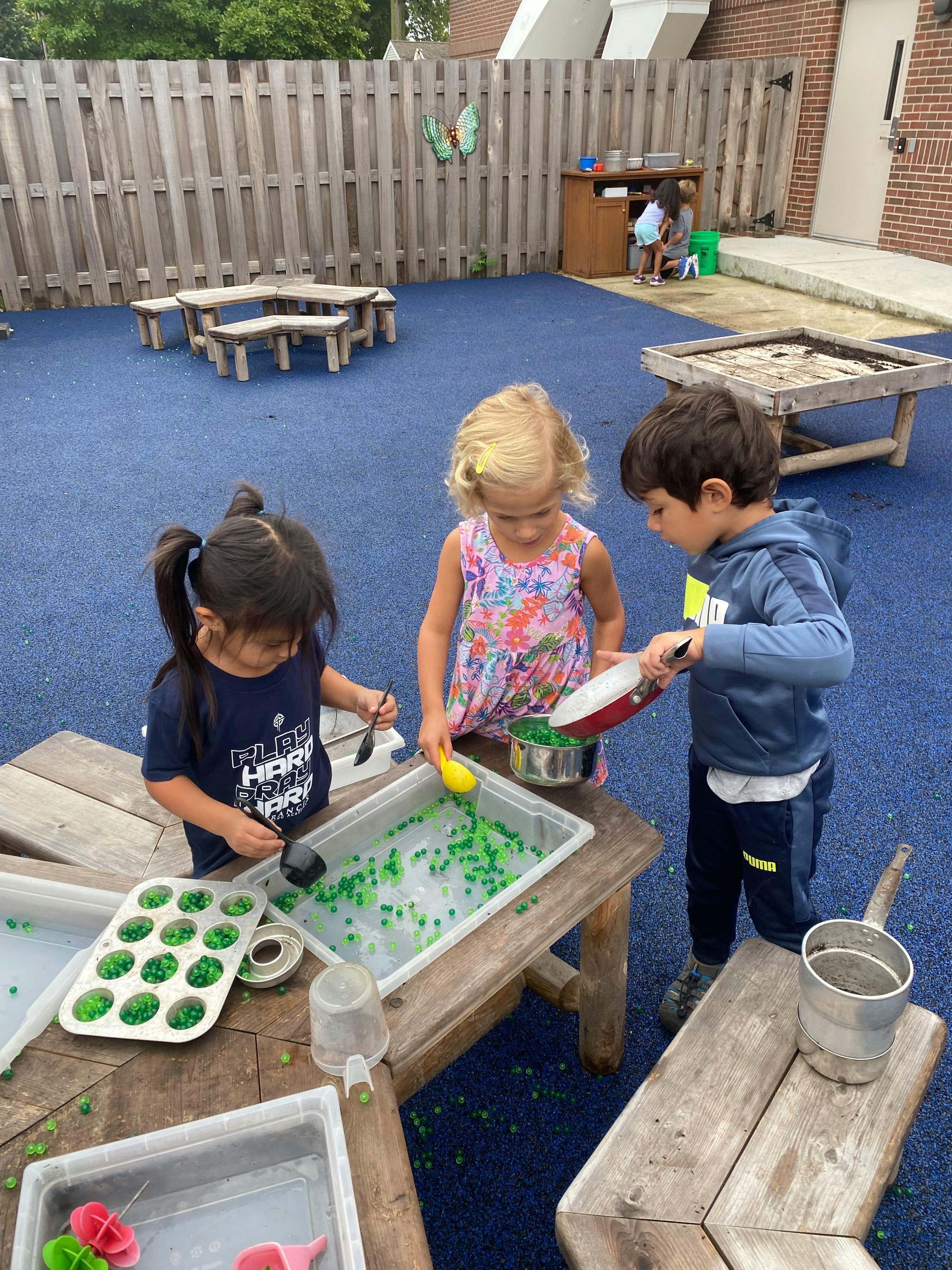 Three children playing with water and green objects at wooden tables outdoors