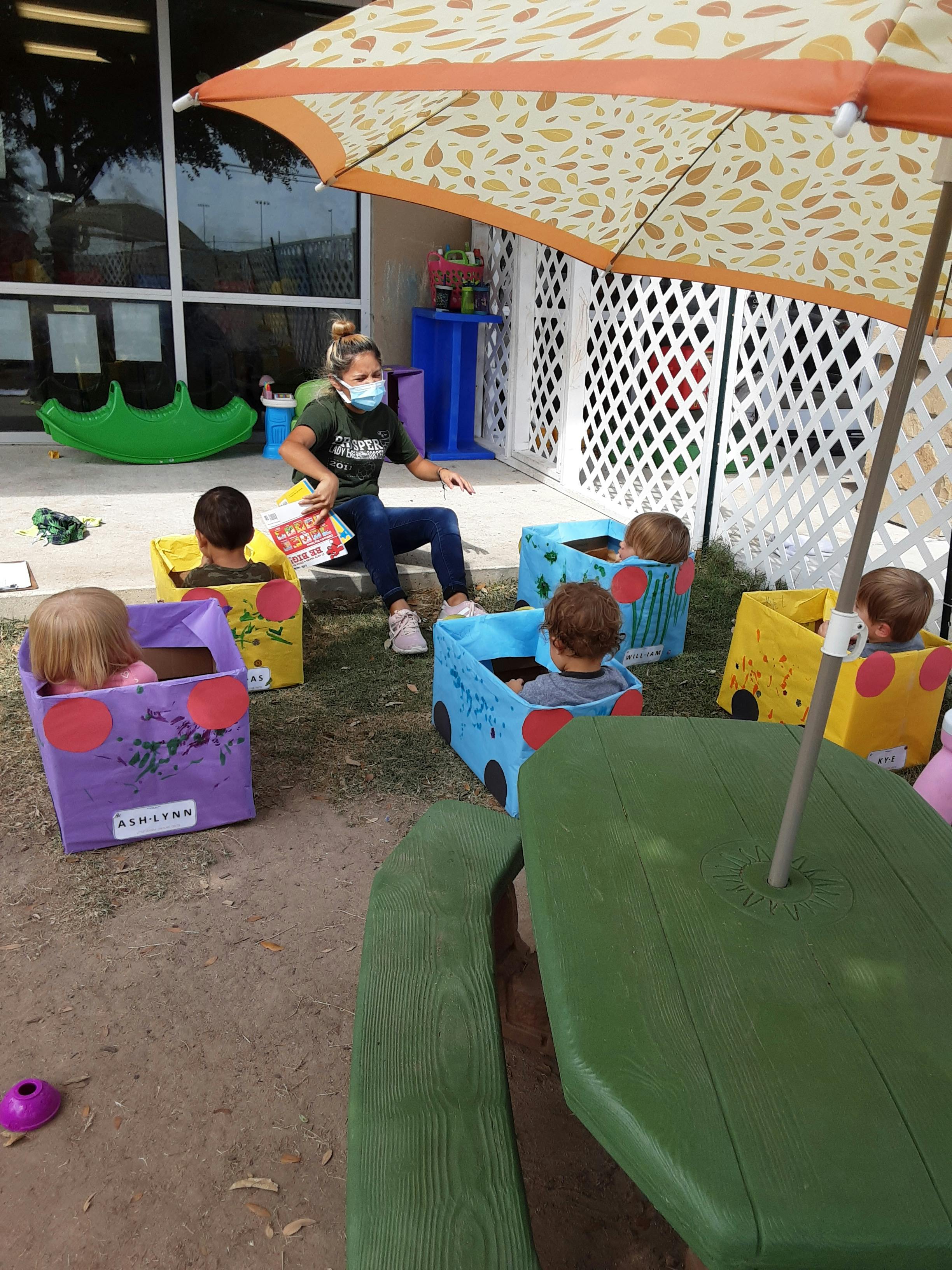 Person in mask reading to children in cardboard cars outdoors
