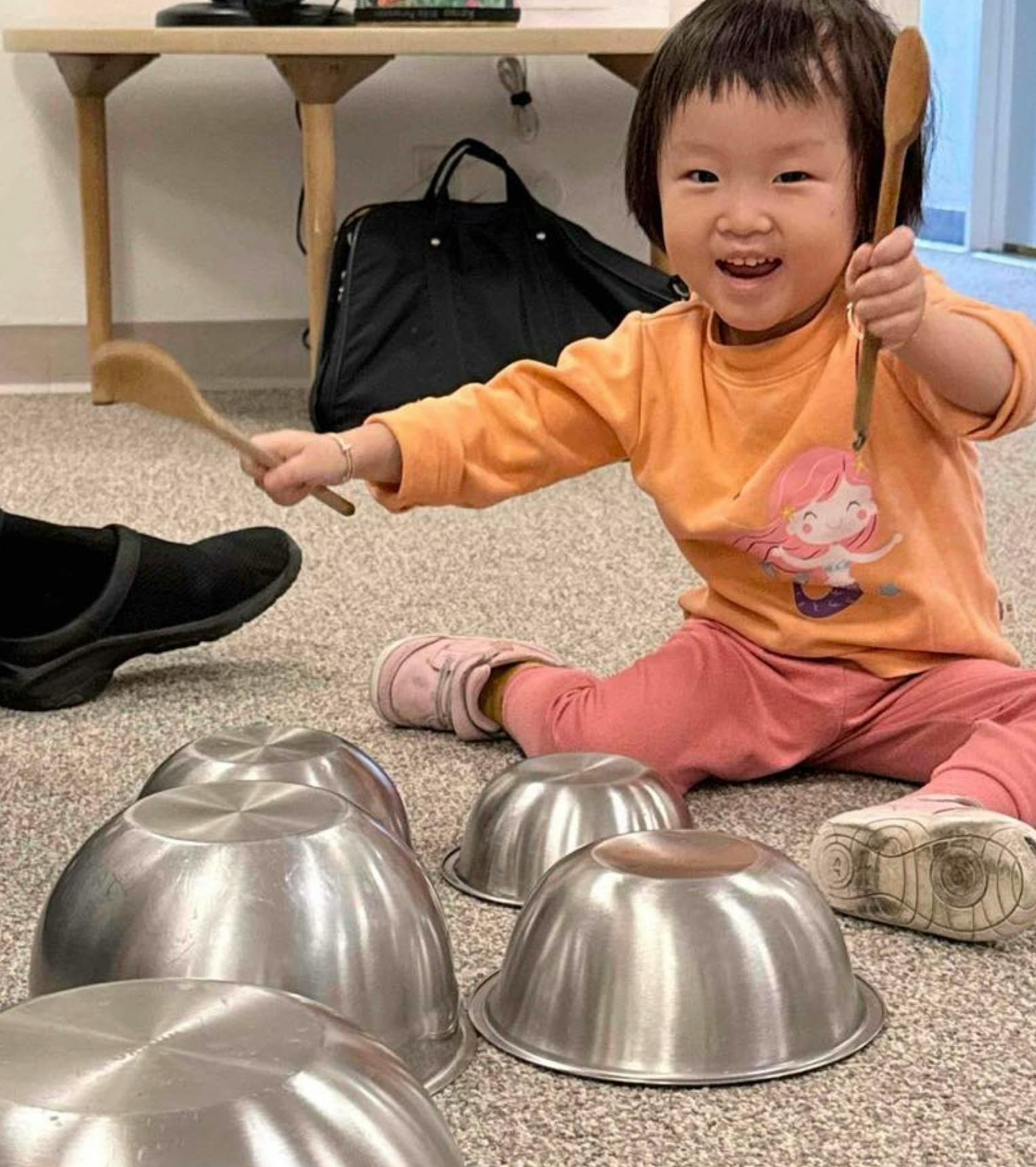 Child sitting on the floor playing with metal bowls using wooden spoons
