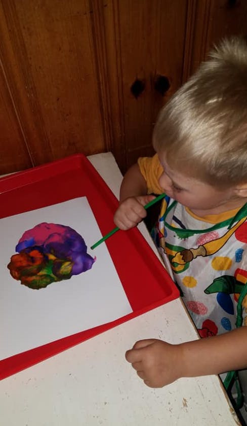 Child blowing paint with a straw on paper in a red tray