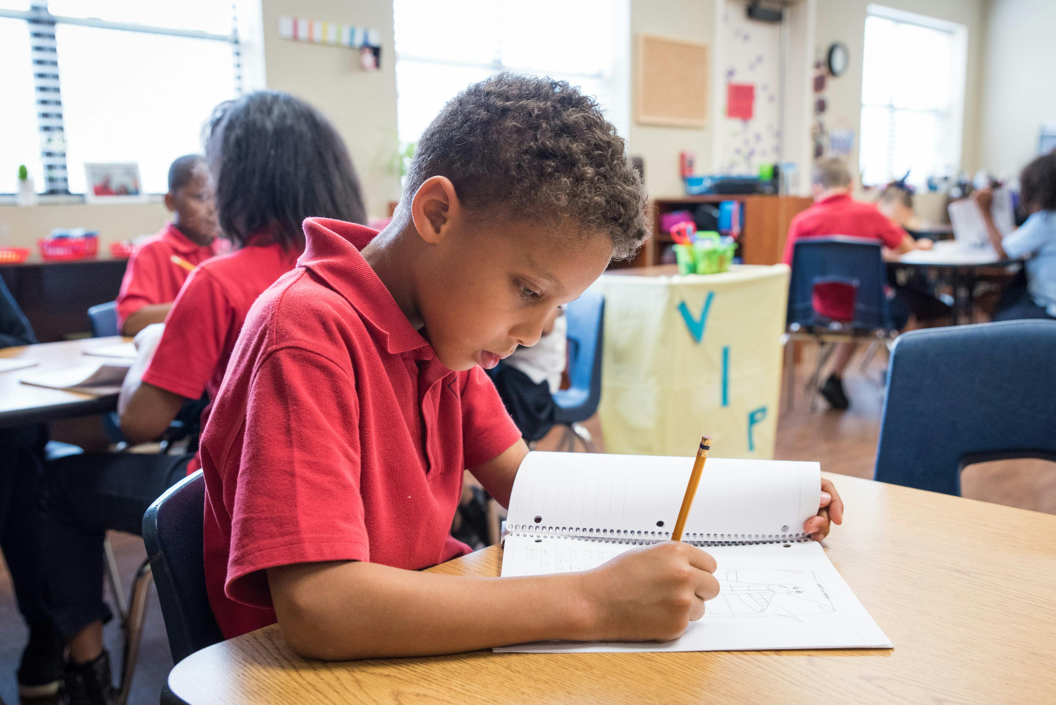 Child writing in a notebook with a pencil in a classroom