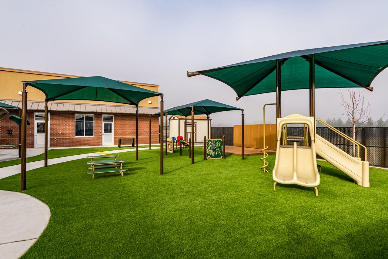 Outdoor playground with green turf, shaded areas, a slide, and musical play equipment