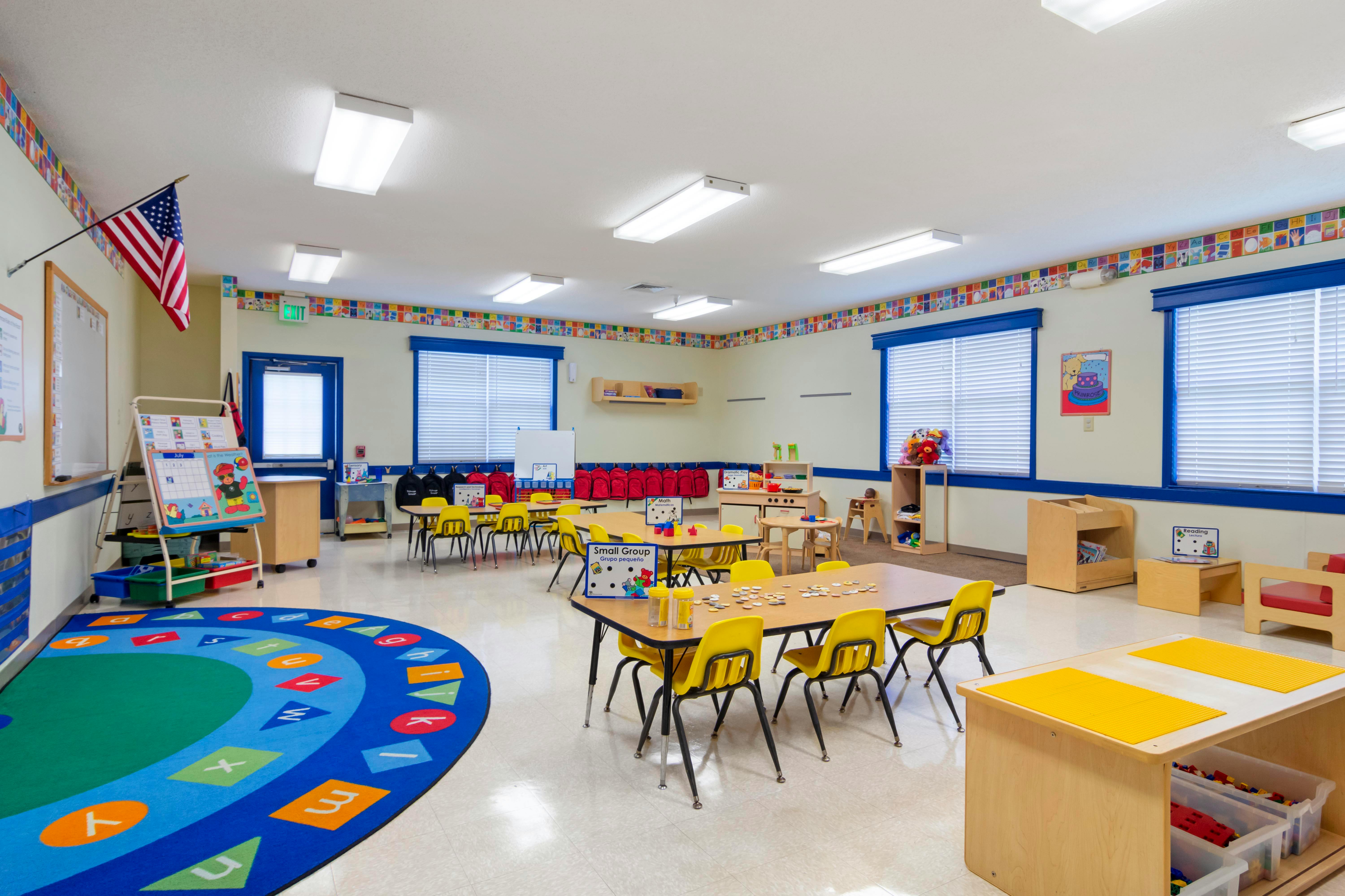 Empty classroom with small tables and chairs, a colorful rug, and an American flag