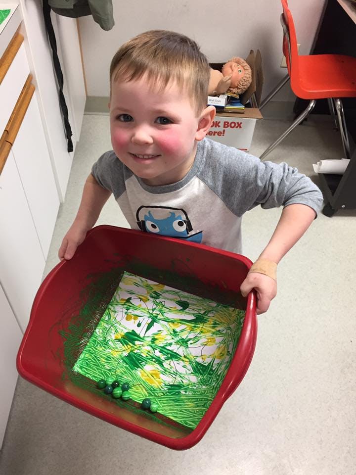 Child holding a red bin with a painting and green marbles inside
