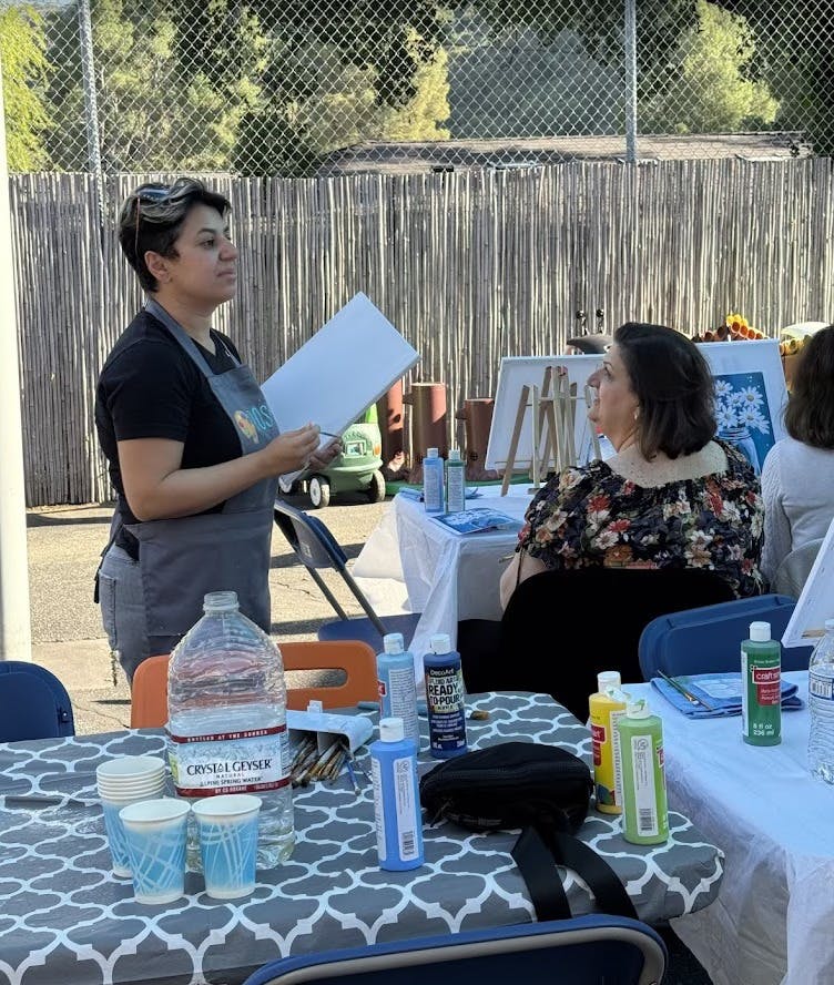 Person in apron holding a canvas, another person seated at a table with art supplies