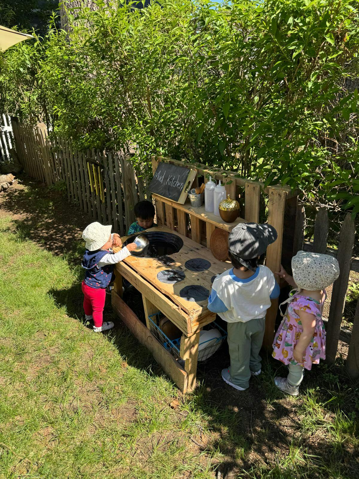 Several children playing at a wooden outdoor mud kitchen with text "Mud Kitchen"