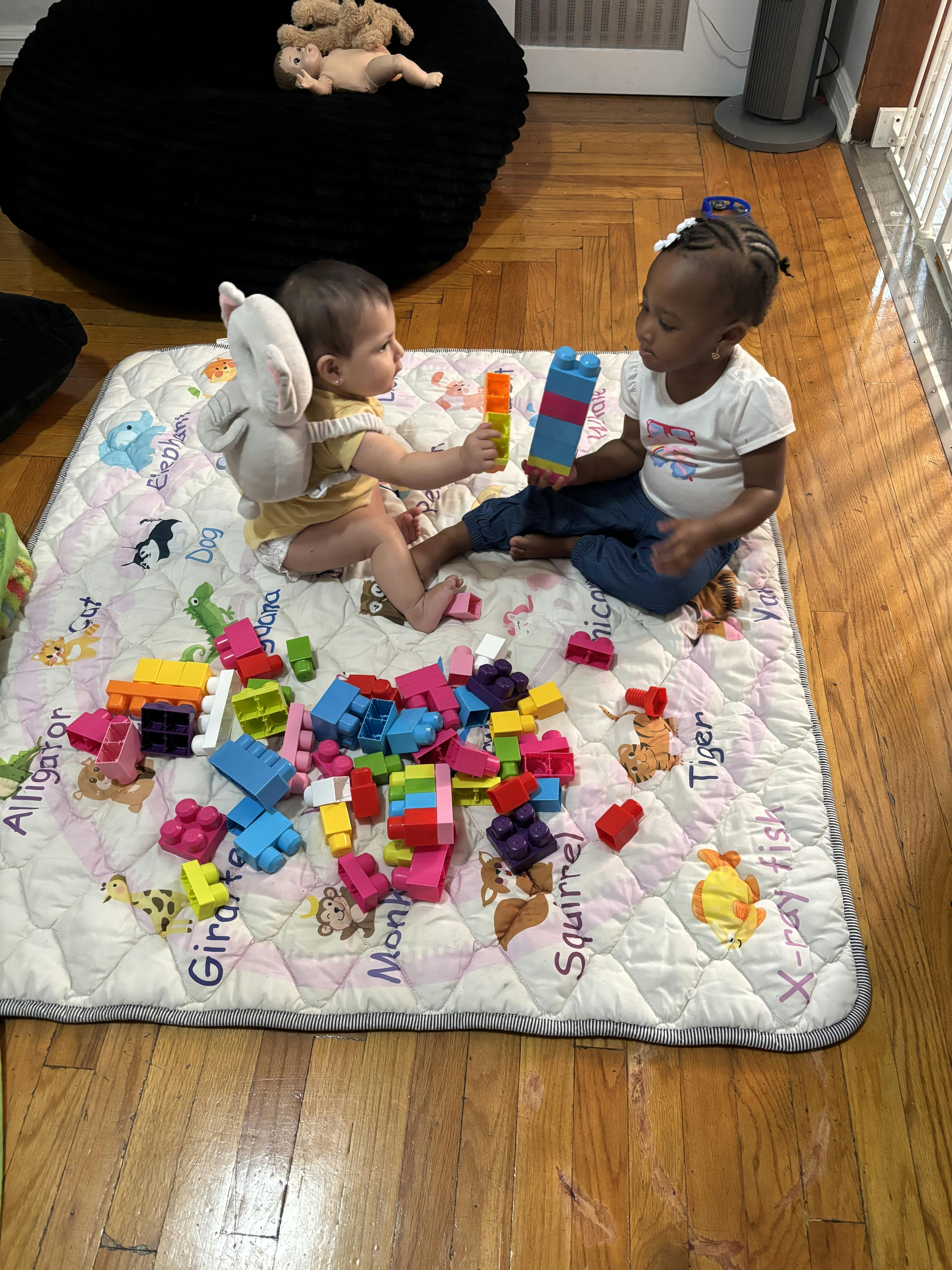 Two children sitting on a floor mat playing with colorful plastic building blocks