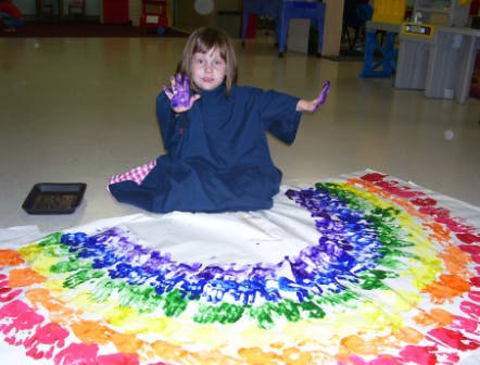 Child with paint on hands sitting next to a rainbow handprint painting