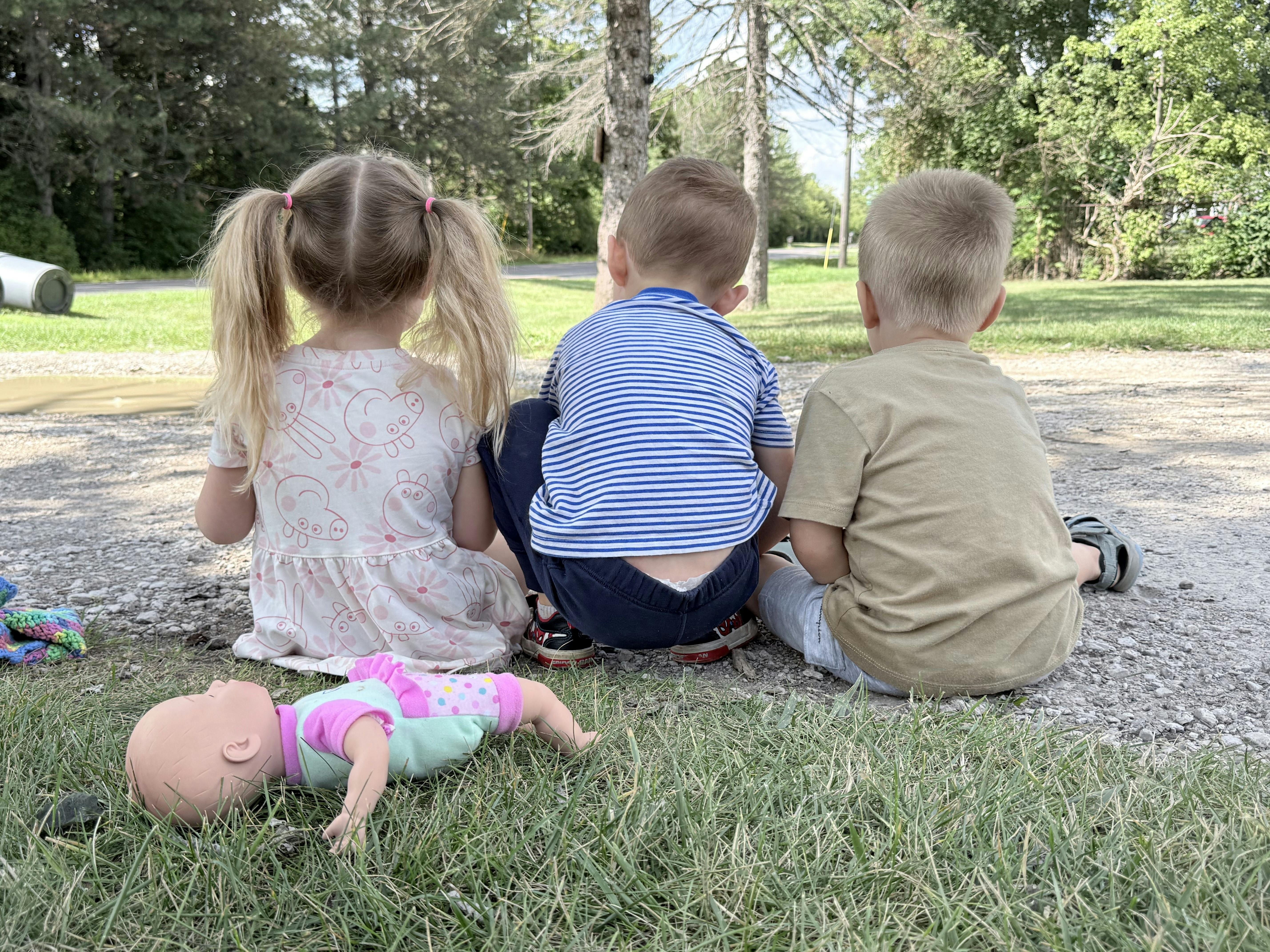 Three children sitting on a gravel path with a doll nearby