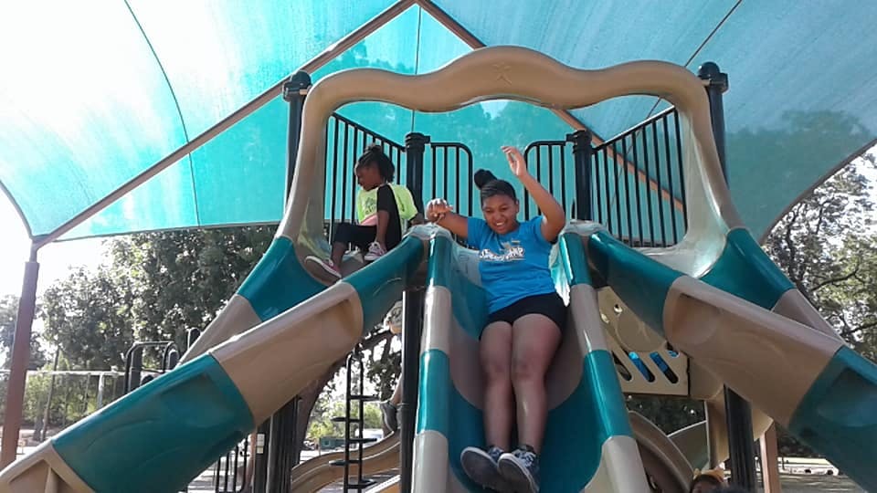 Teen on a slide with arms raised, child sitting on playground equipment