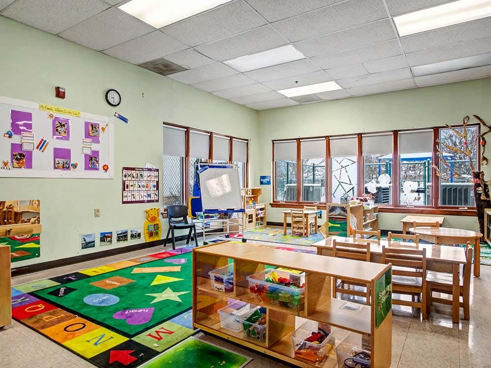Classroom with small tables and chairs, a colorful alphabet rug, and large windows