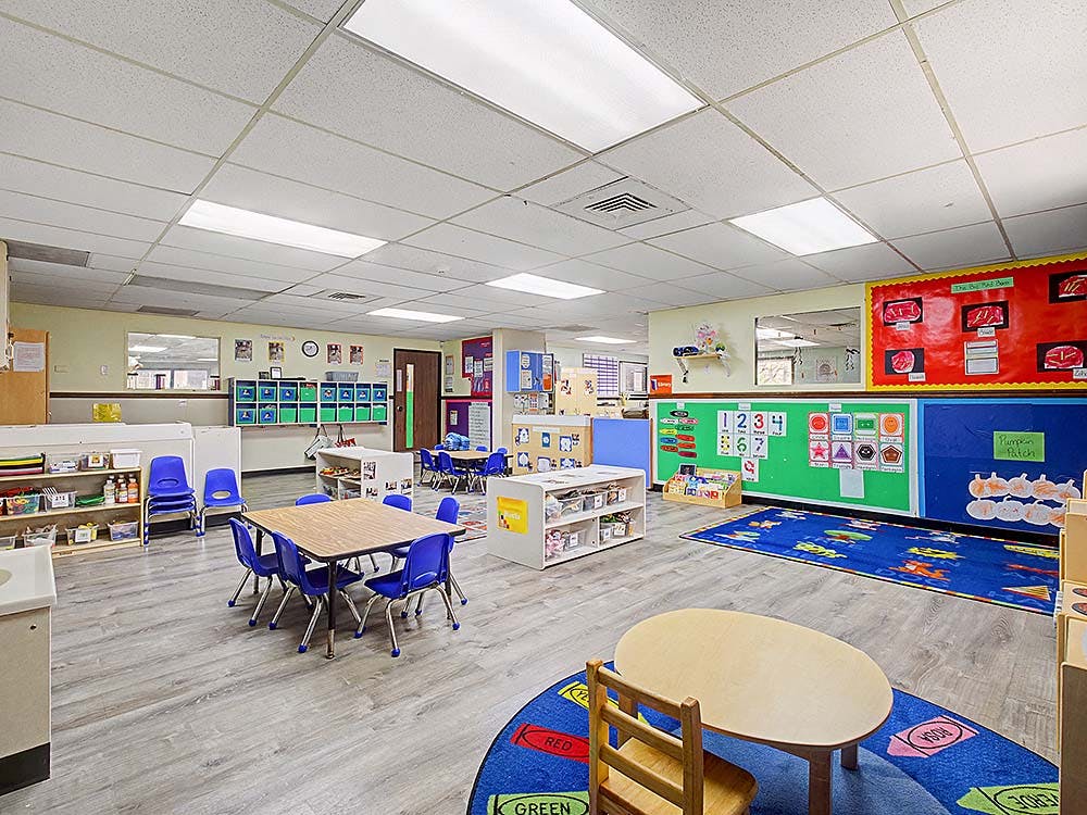 Empty classroom with small tables and chairs, colorful rugs, and learning charts on walls