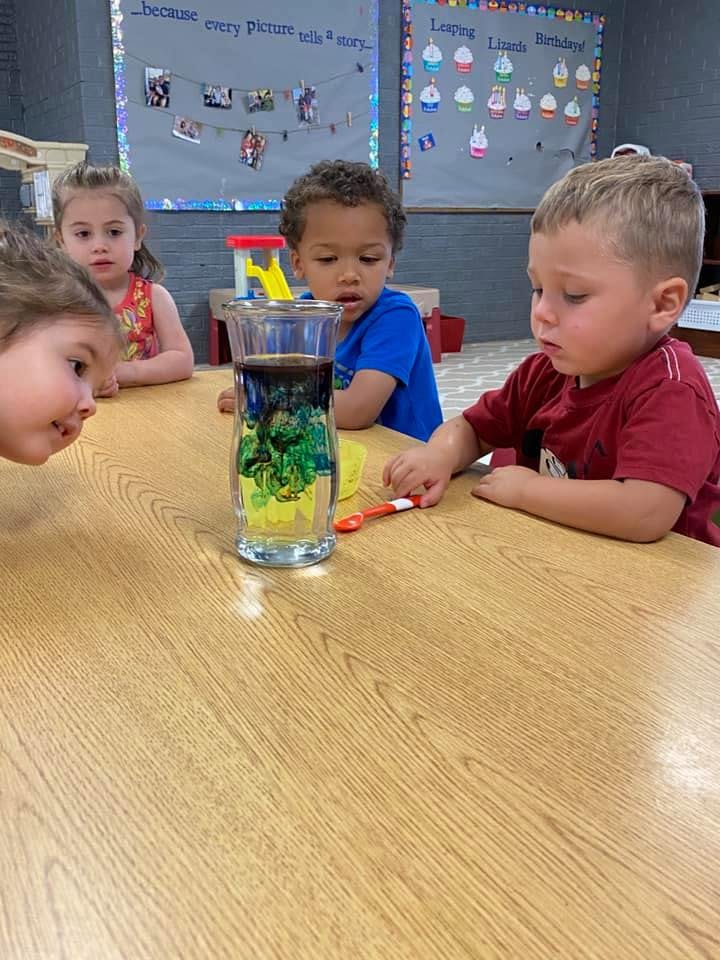 Three children at a table looking at a glass with liquid and green substance