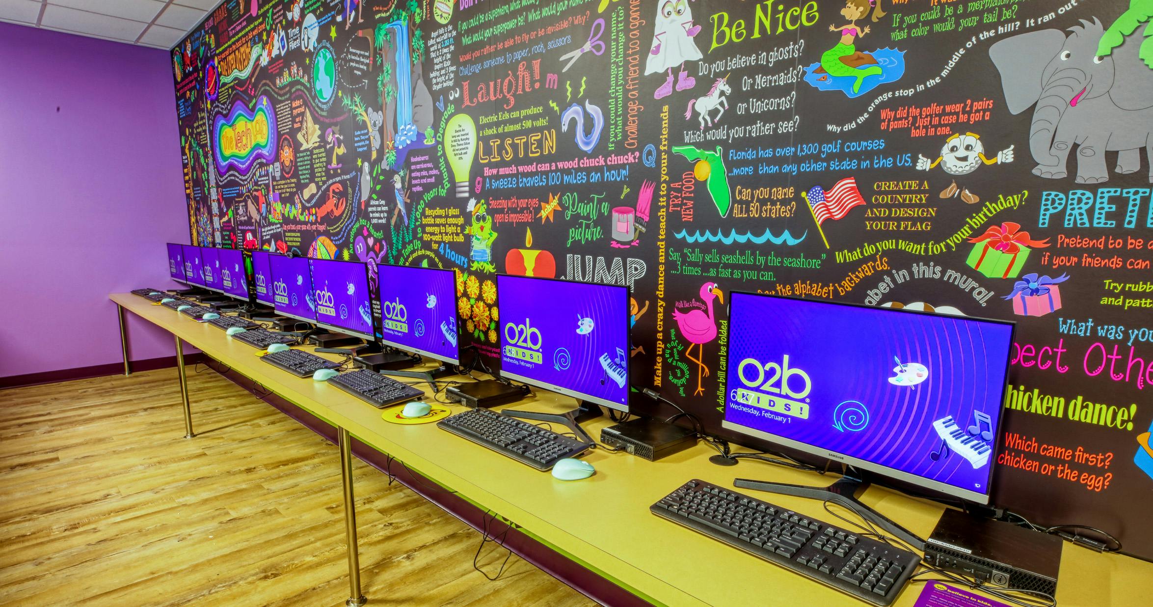 A line of computer monitors and keyboards on a long table in front of a colorful mural