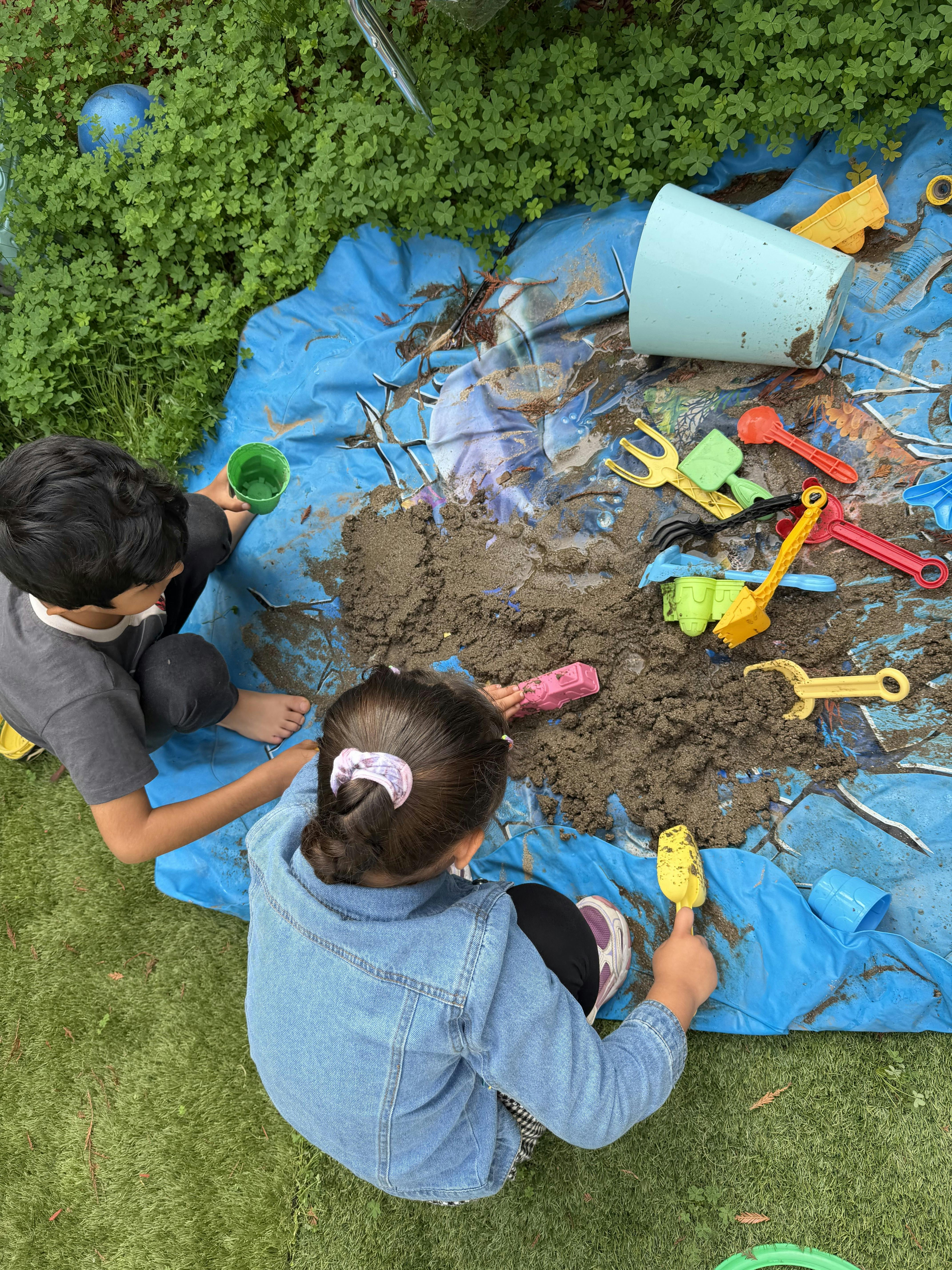 Two children playing with sand and plastic tools on a blue tarp outdoors