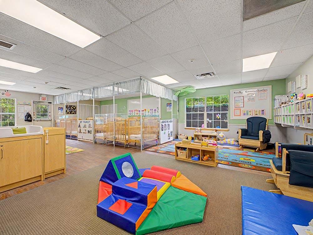 Classroom with cribs, colorful play mat, shelves, and seating