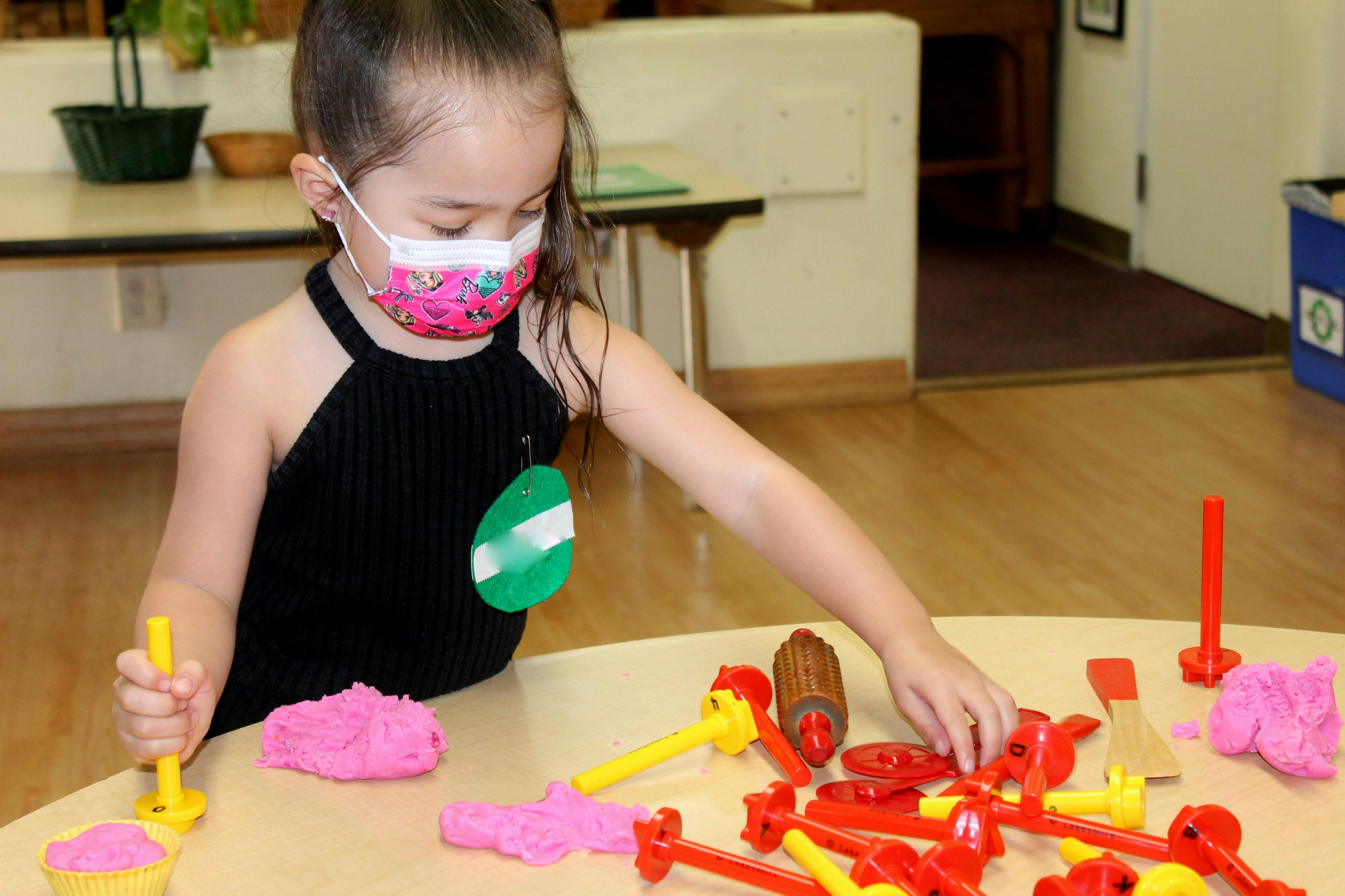 Child in a mask playing with pink dough and red and yellow toys