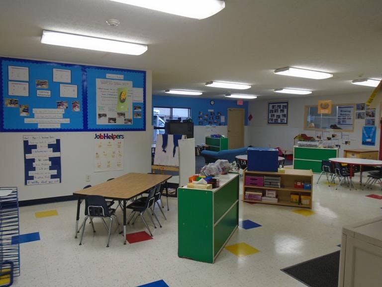 An empty classroom with small tables and chairs, bulletin boards, and storage units