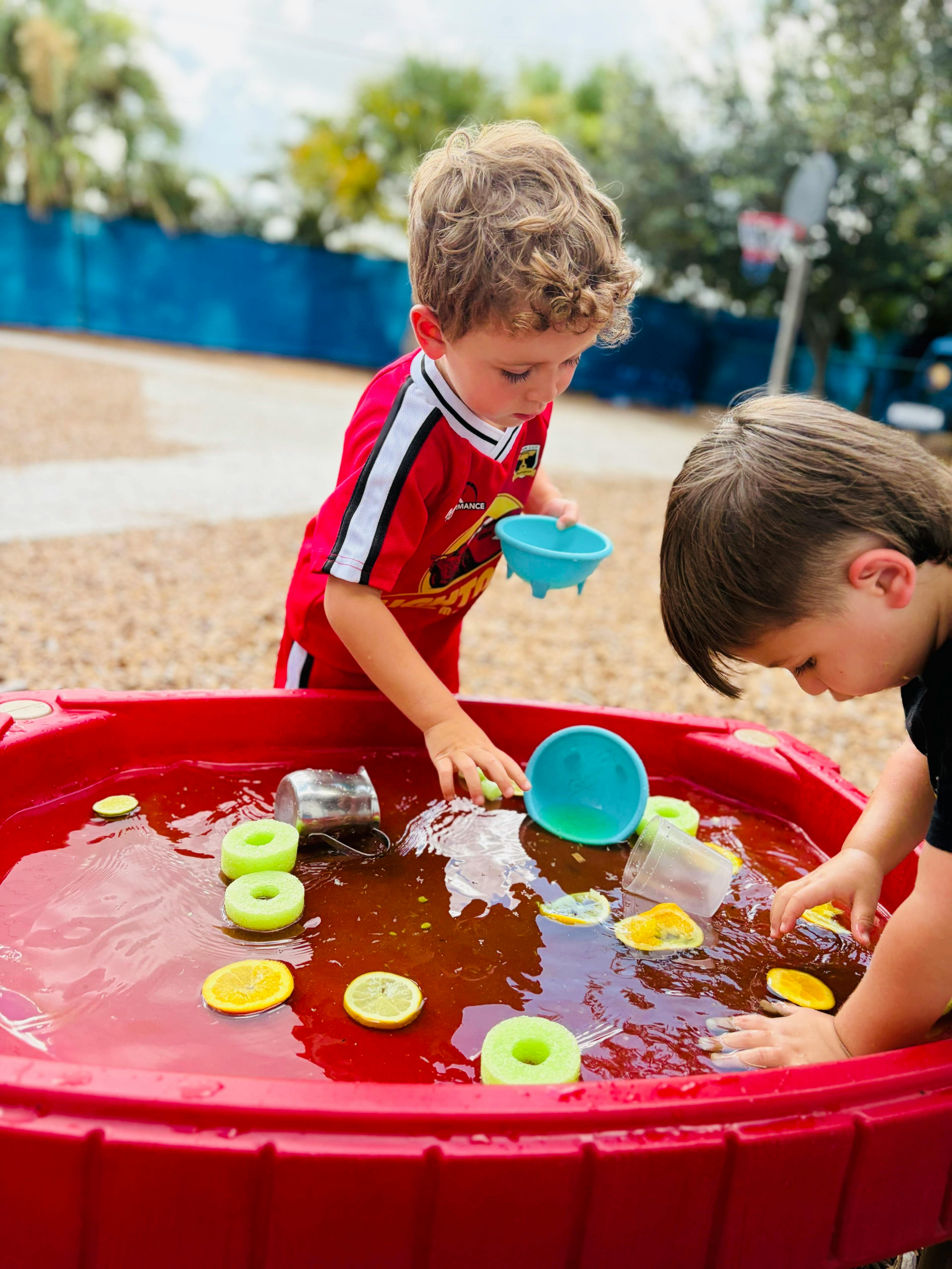 Two children playing with water and fruit slices in a red plastic bin