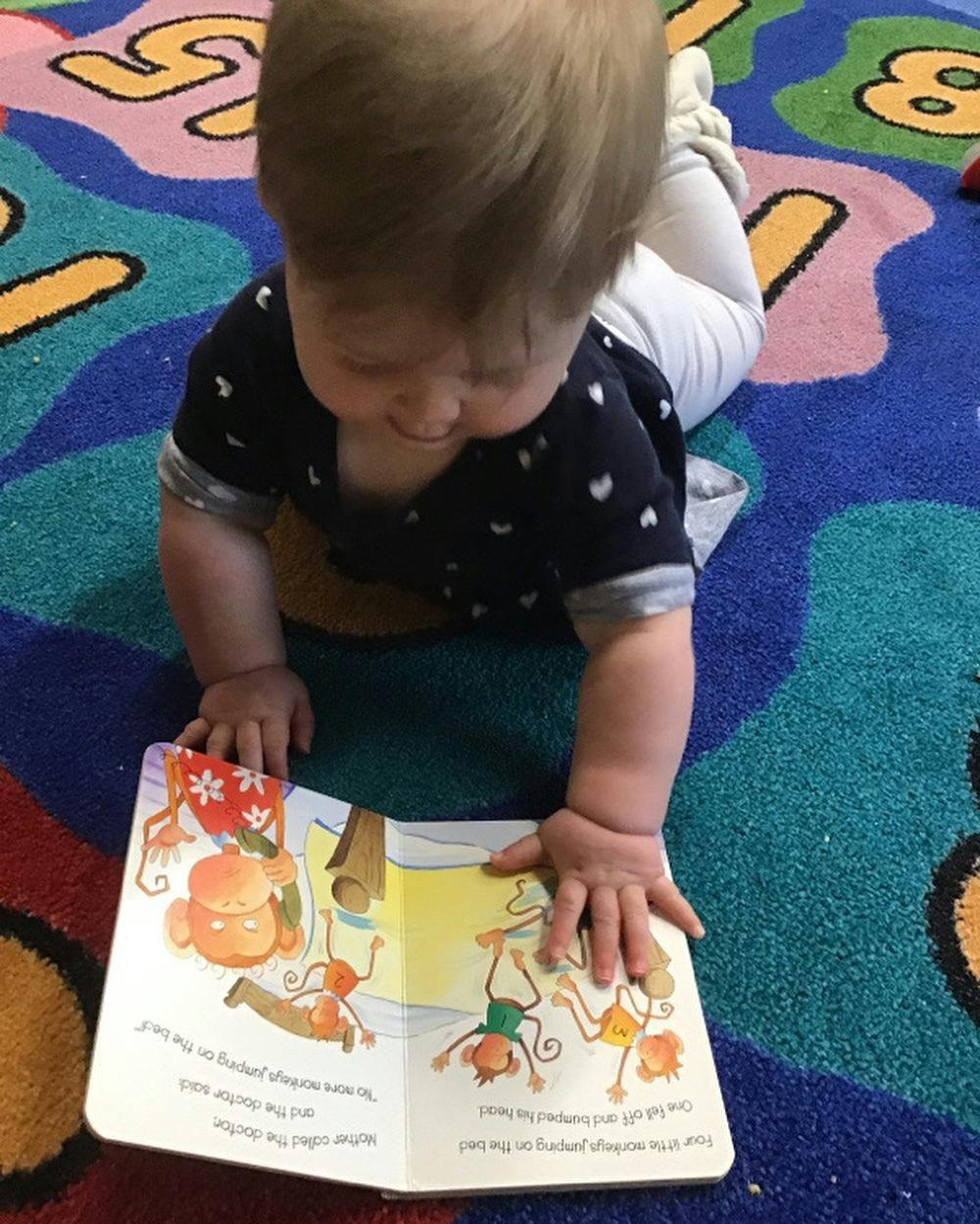 Child on stomach looking at an open book on a colorful rug