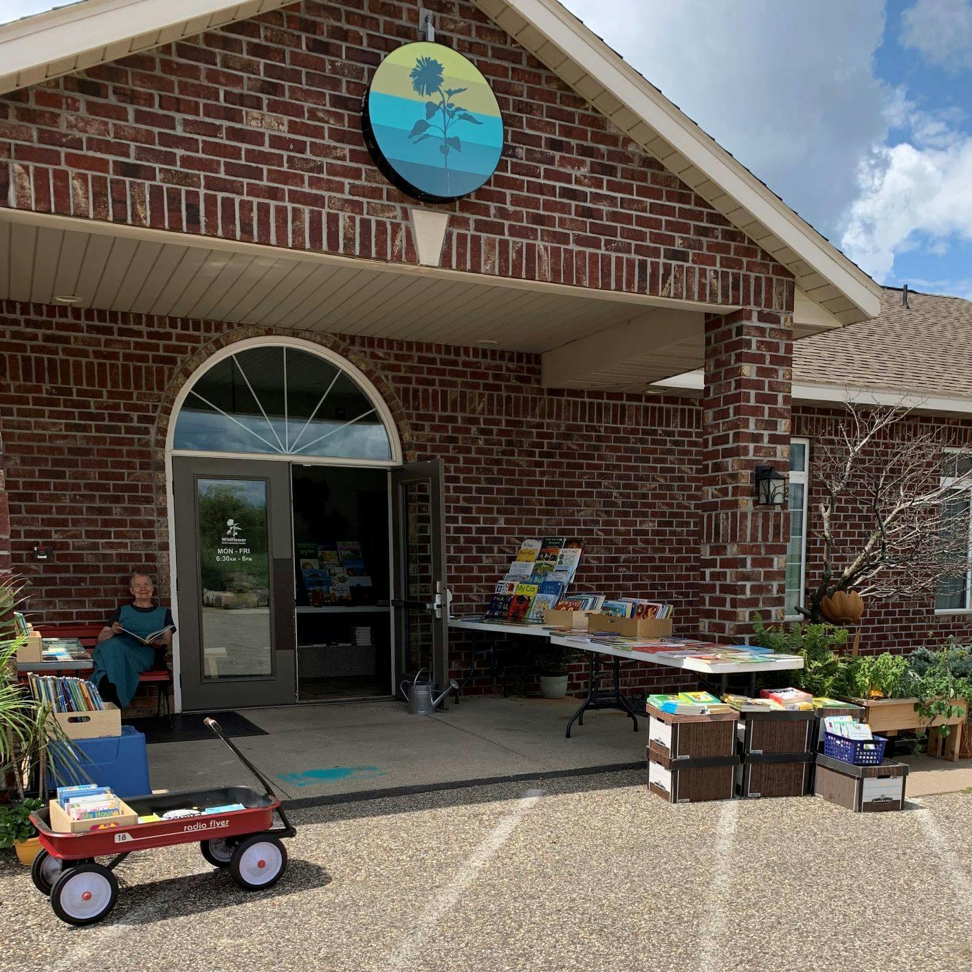 Person sitting outside a brick building with tables of items and a red wagon