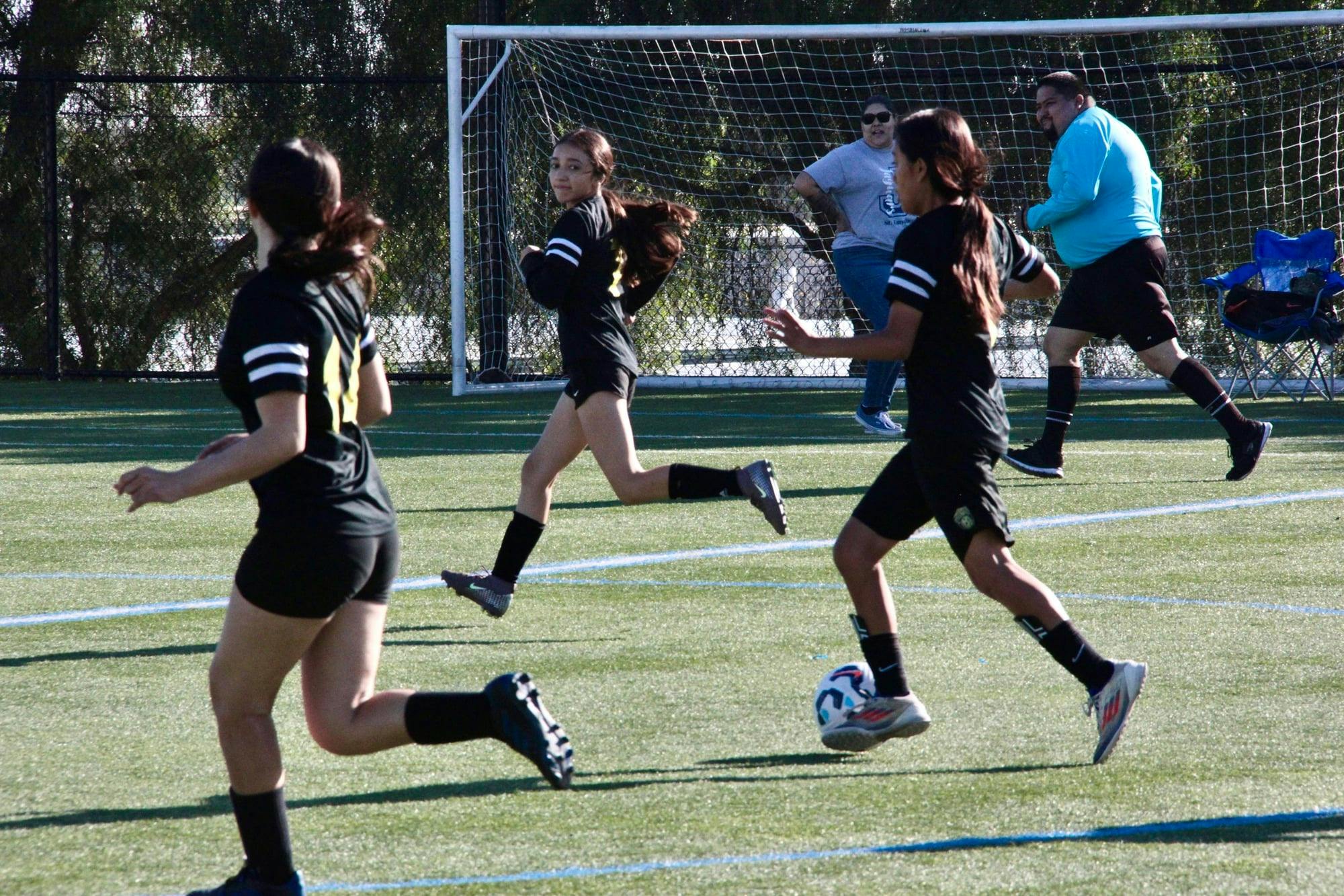 Three people in black soccer uniforms running on a field with a soccer ball