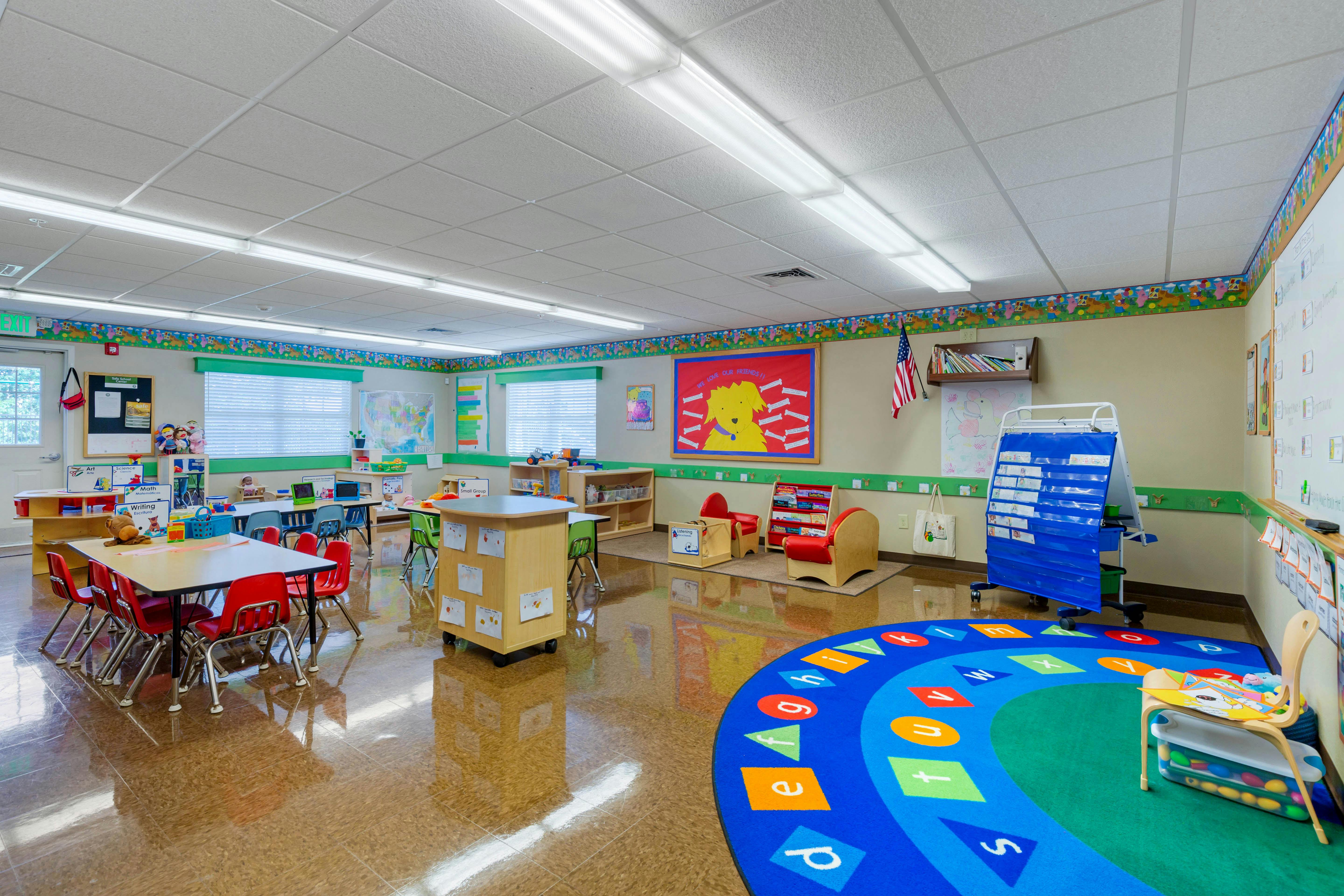 Empty classroom with small tables and chairs, and a colorful alphabet rug