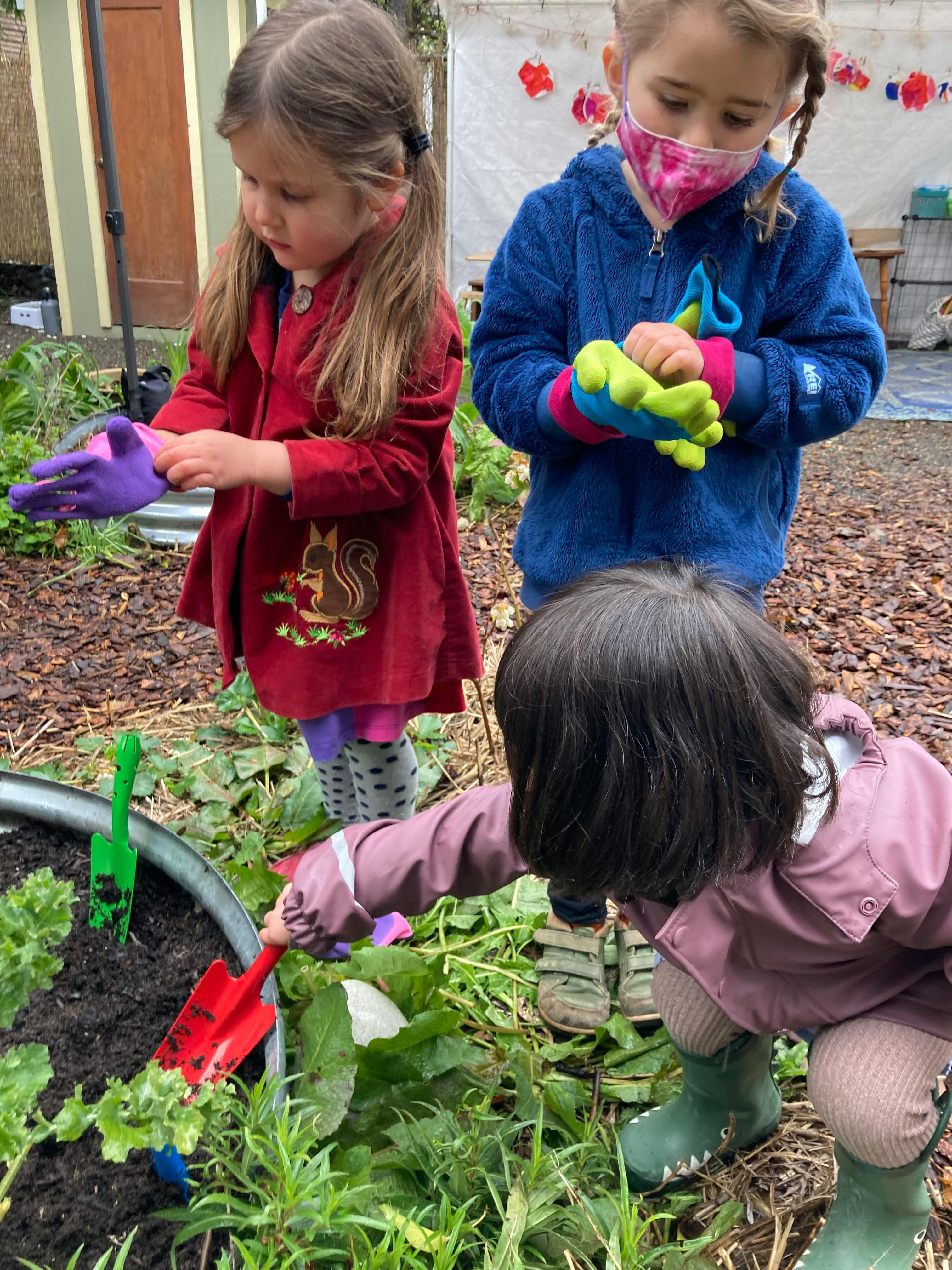 Three children gardening, one holding a trowel, another wearing gloves