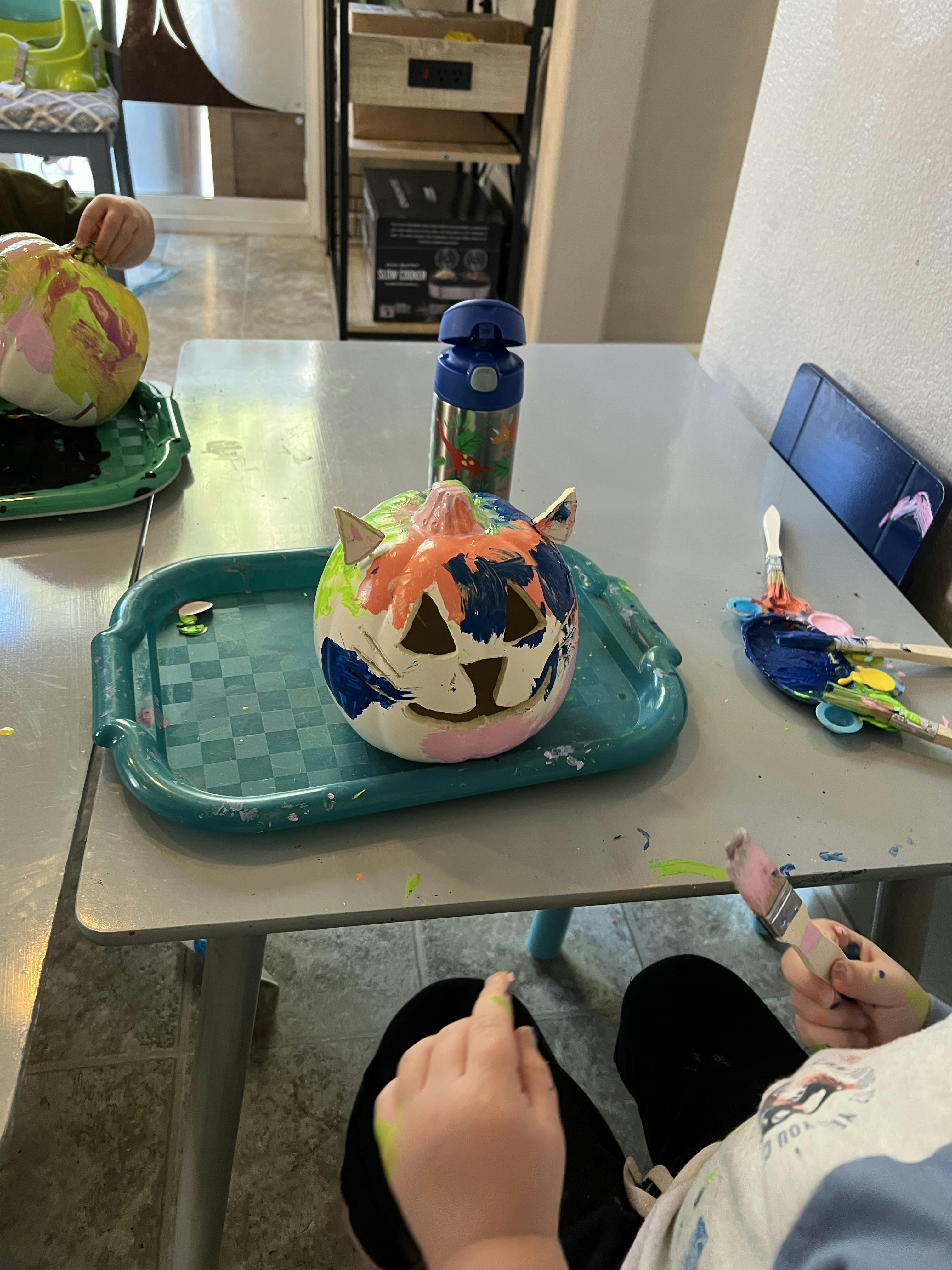 Child painting a carved pumpkin with cat ears, another pumpkin on the table