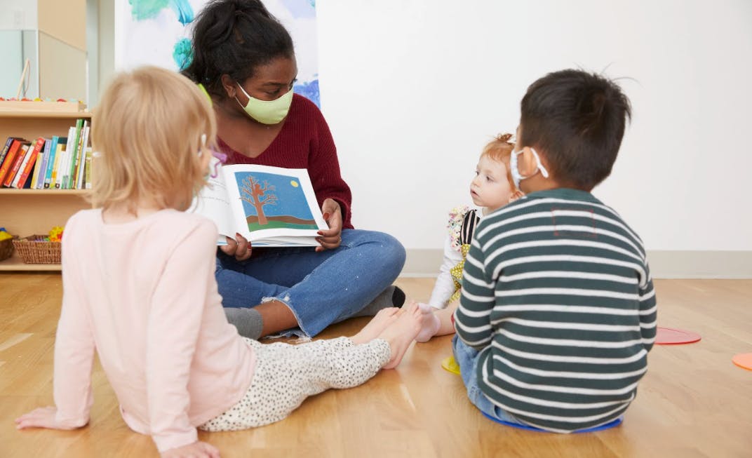 Person in mask reading a book to three children in a classroom