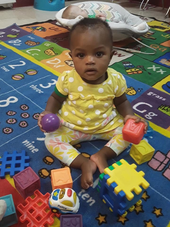 Child sitting on a playmat with toys, holding a purple ball and a red block