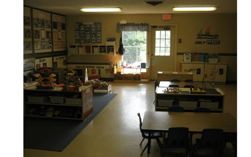 An empty classroom with small tables and chairs, shelves with toys