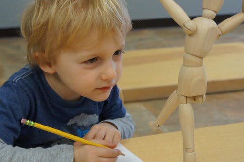 Child drawing with a pencil, looking at a wooden art mannequin