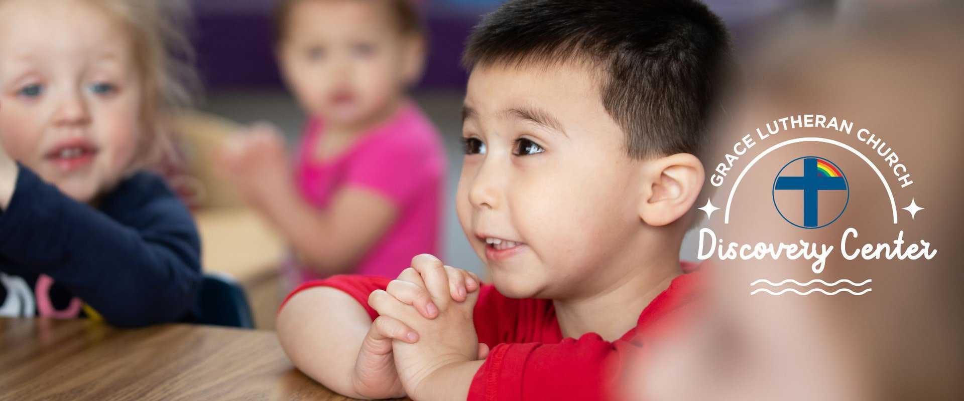 Child with hands clasped, looking up, with other children in background