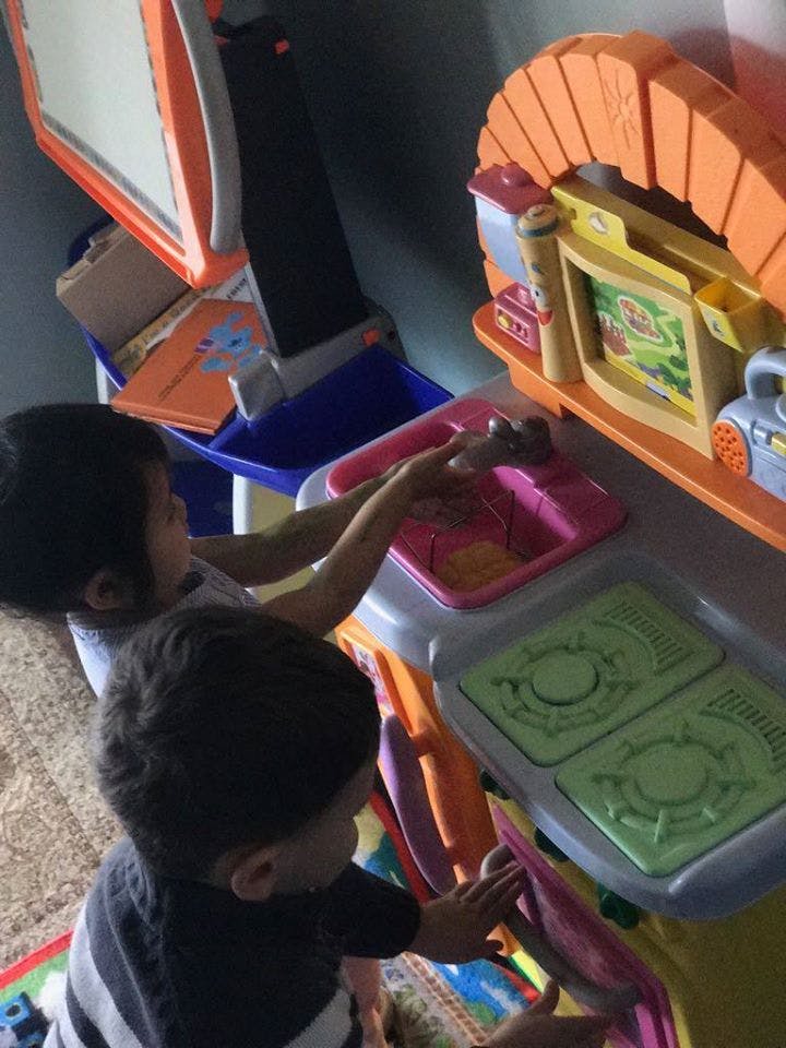 Two children playing with a toy kitchen set, one at a sink and the other at a stove