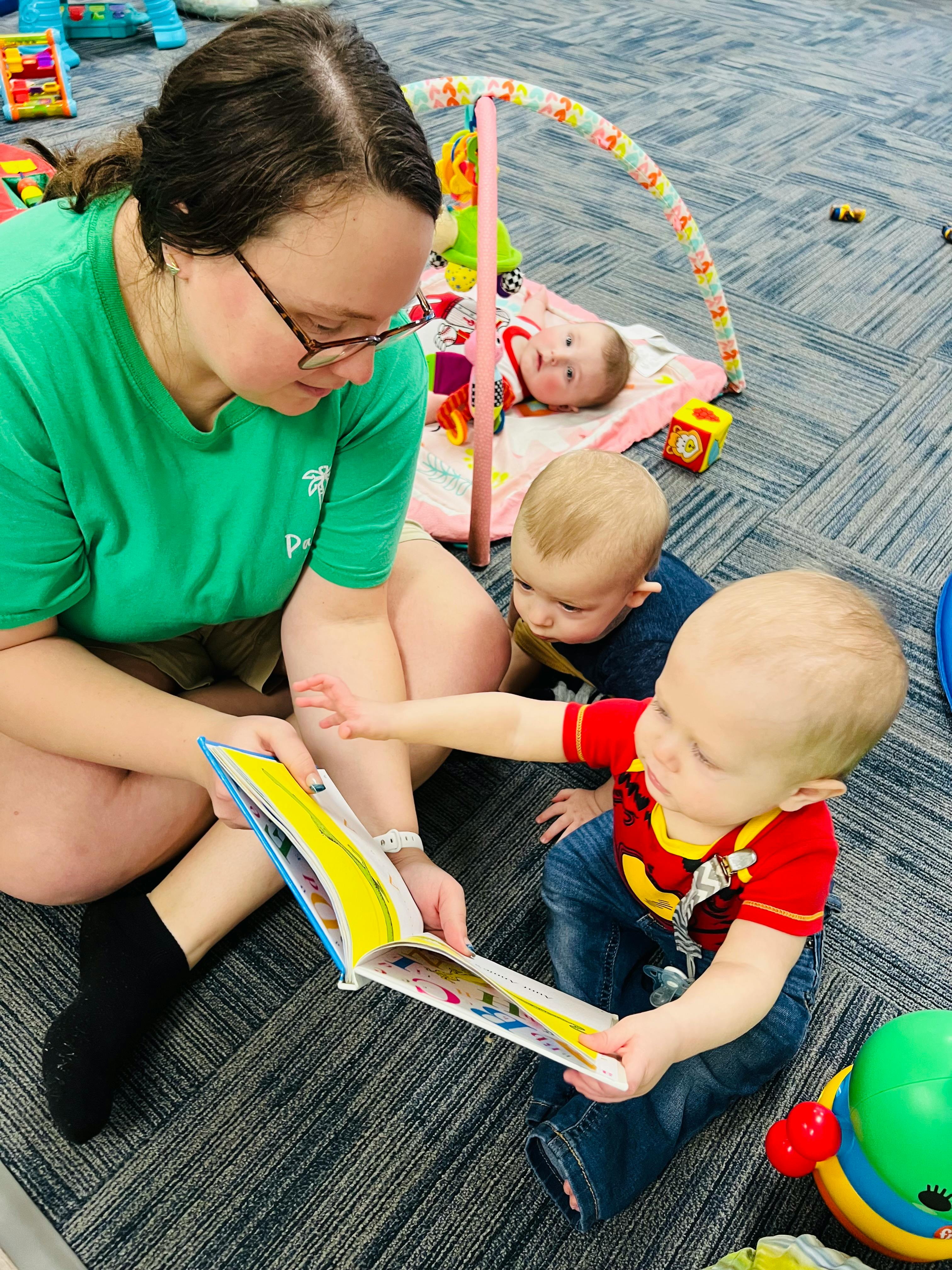 Person reading a book to two children, with another child on a playmat
