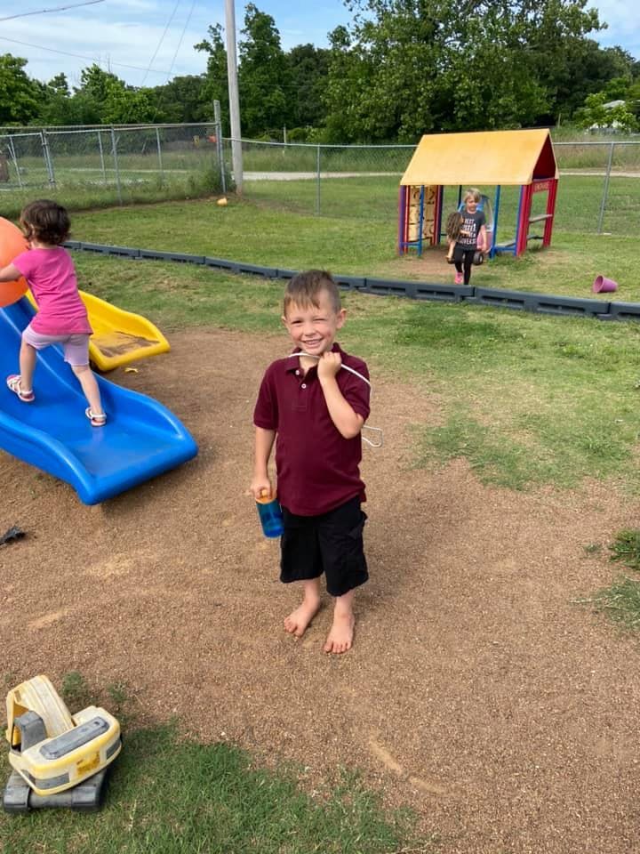 Child standing barefoot on dirt playground, another child on a slide