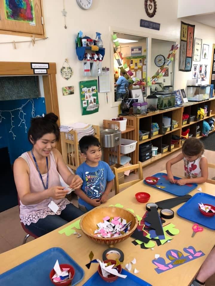 Person and two children doing crafts at a table in a classroom
