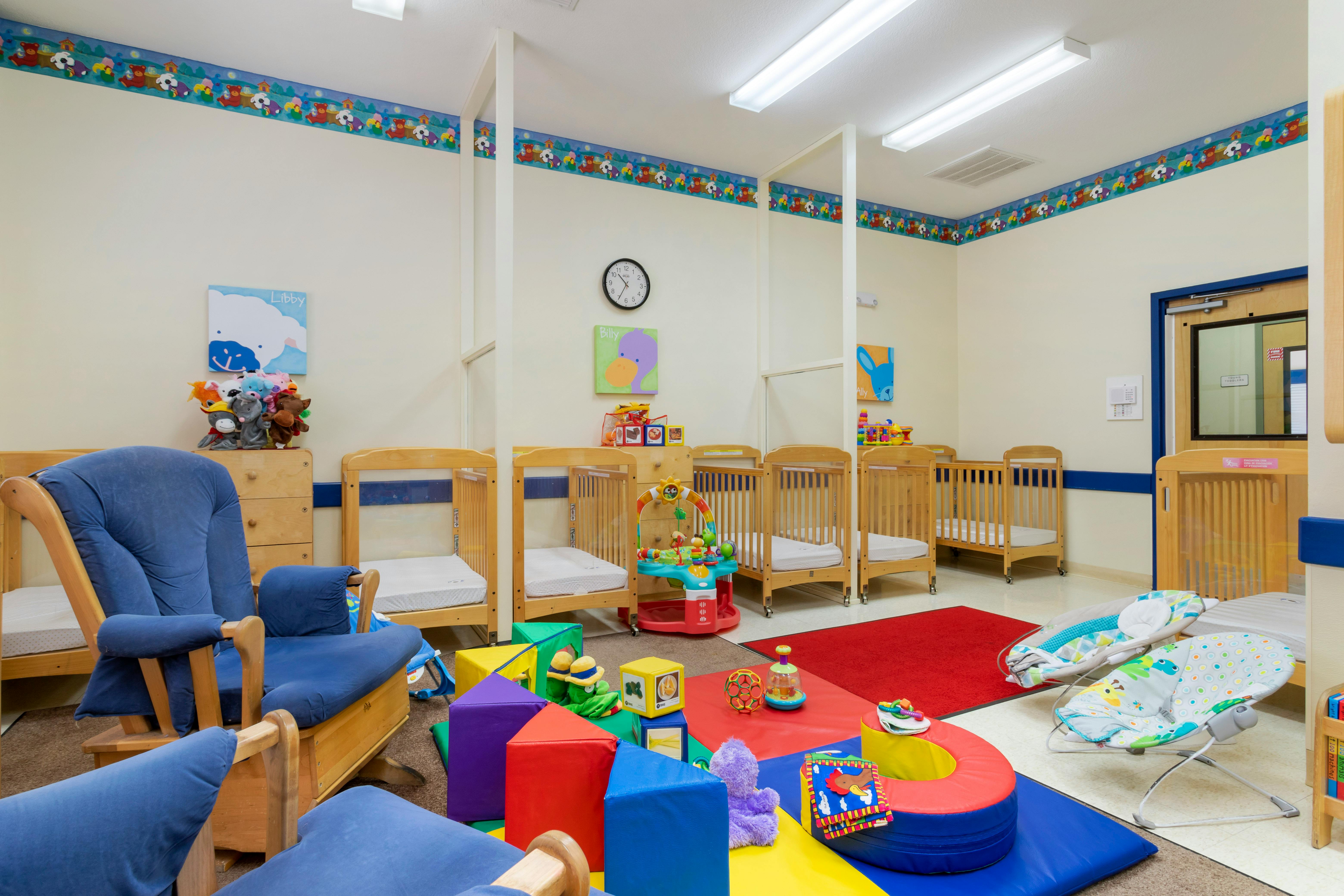 A colorful childcare room with cribs, a rocking chair, and play mats with toys