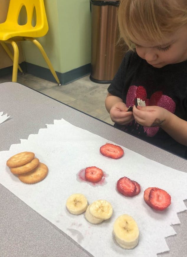 Child peeling a snack wrapper next to crackers, strawberries, and bananas