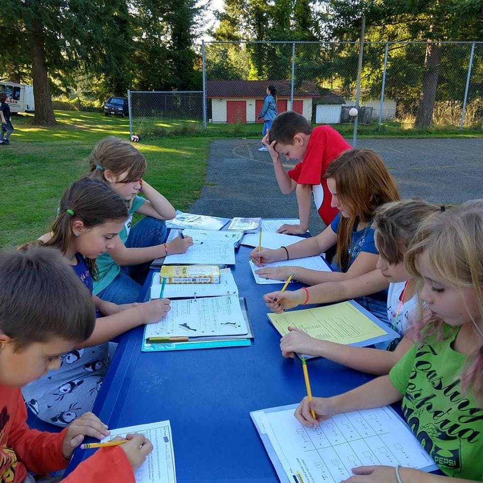 Children and teens writing at a long table outdoors