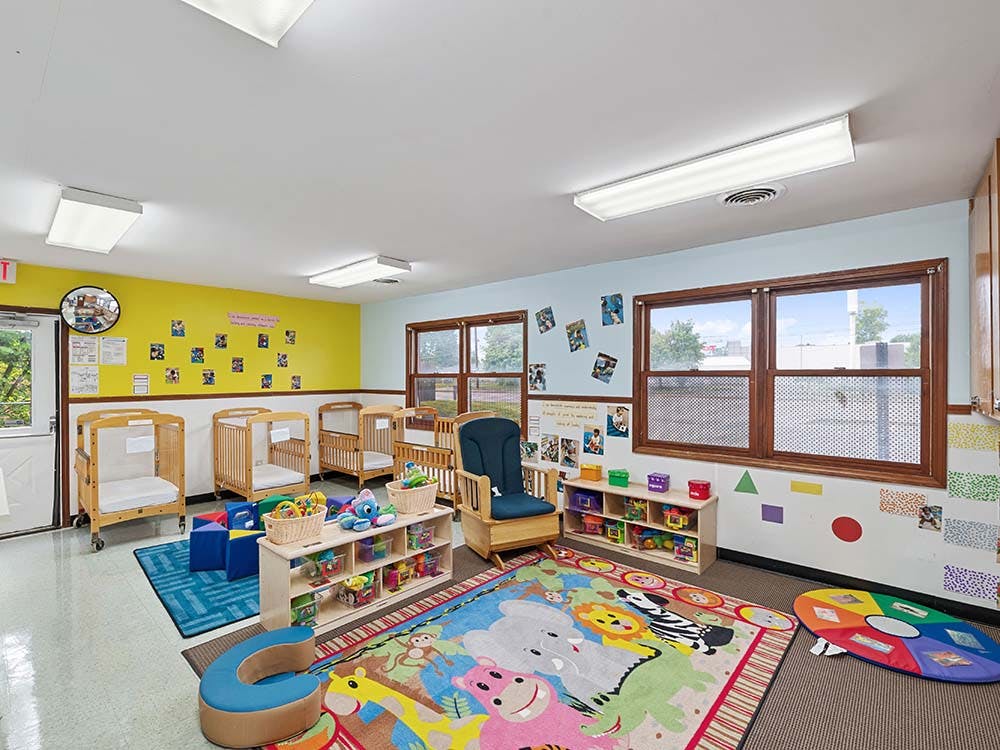 A colorful daycare room with several cribs, a rocking chair, and toys on shelves