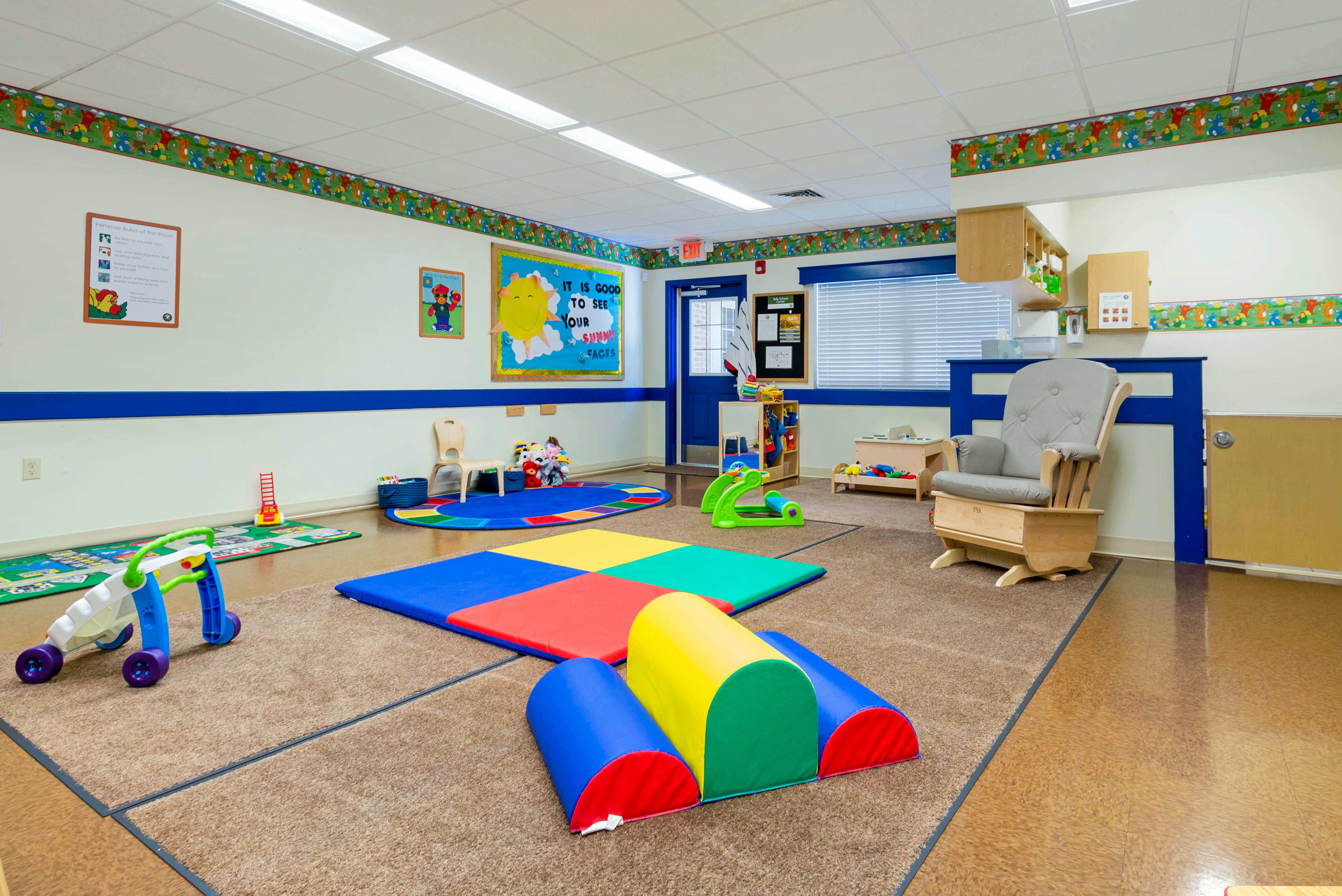 Playroom with colorful mats, a wooden rocking chair, and a bulletin board that says "IT IS GOOD TO SEE YOUR SUNNY FACES"