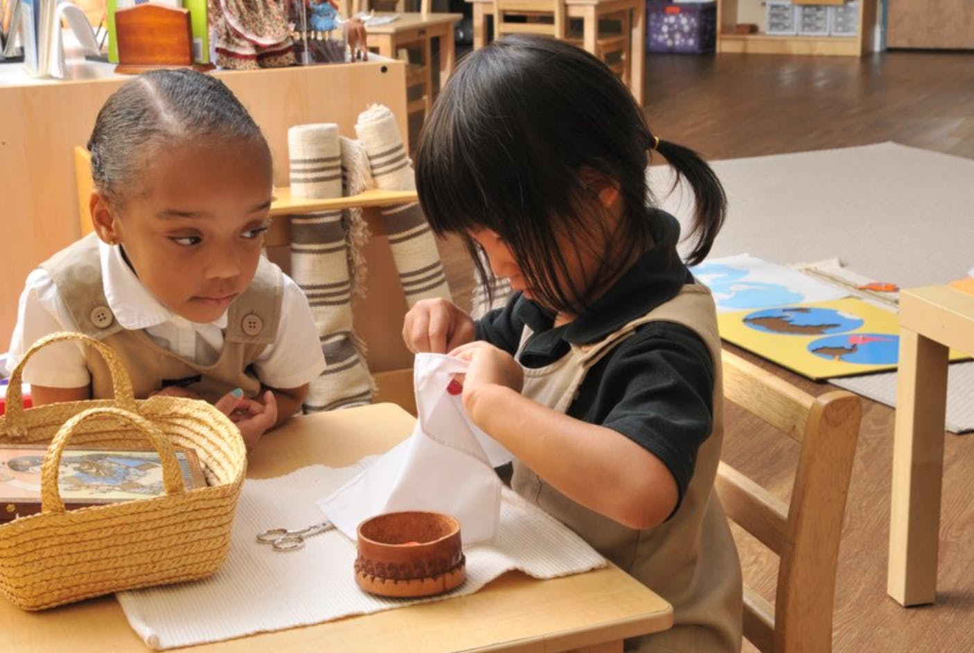 Two children at a table, one sewing and the other watching